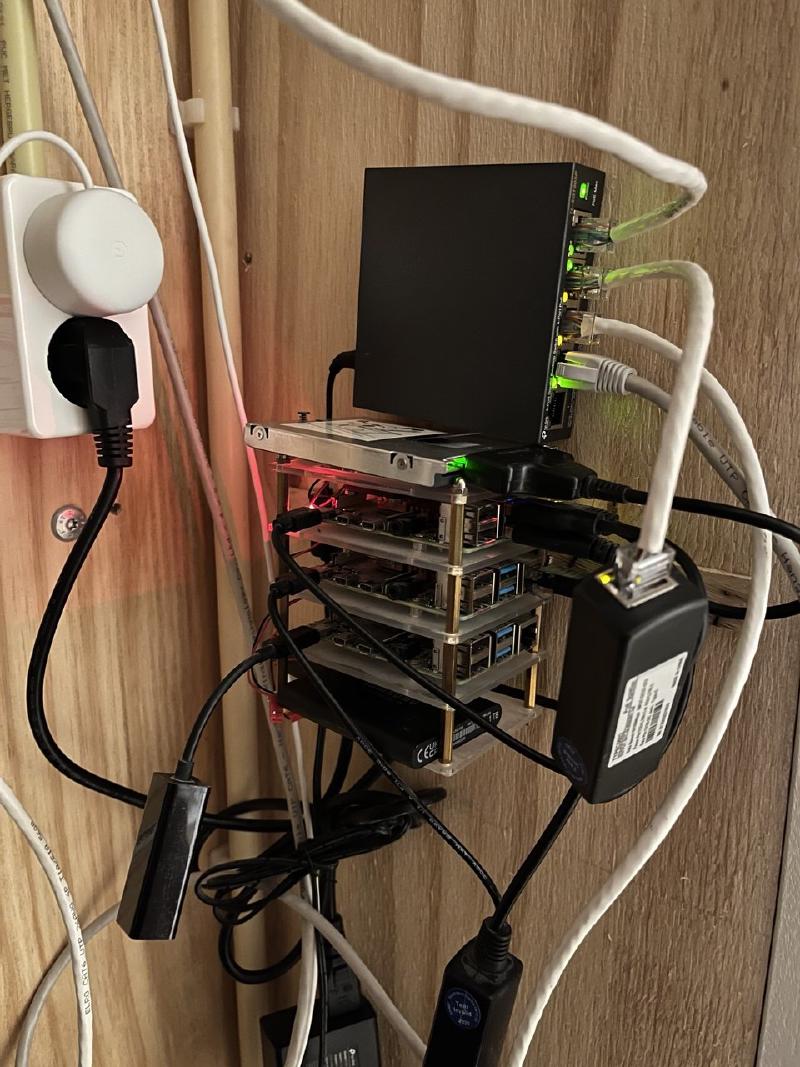 Three Raspberry Pis in a stacked acrylic case with a USB disk on top, a small PoE switch above them, and PoE splitters, mounted on the wooden wall in my server hideout.